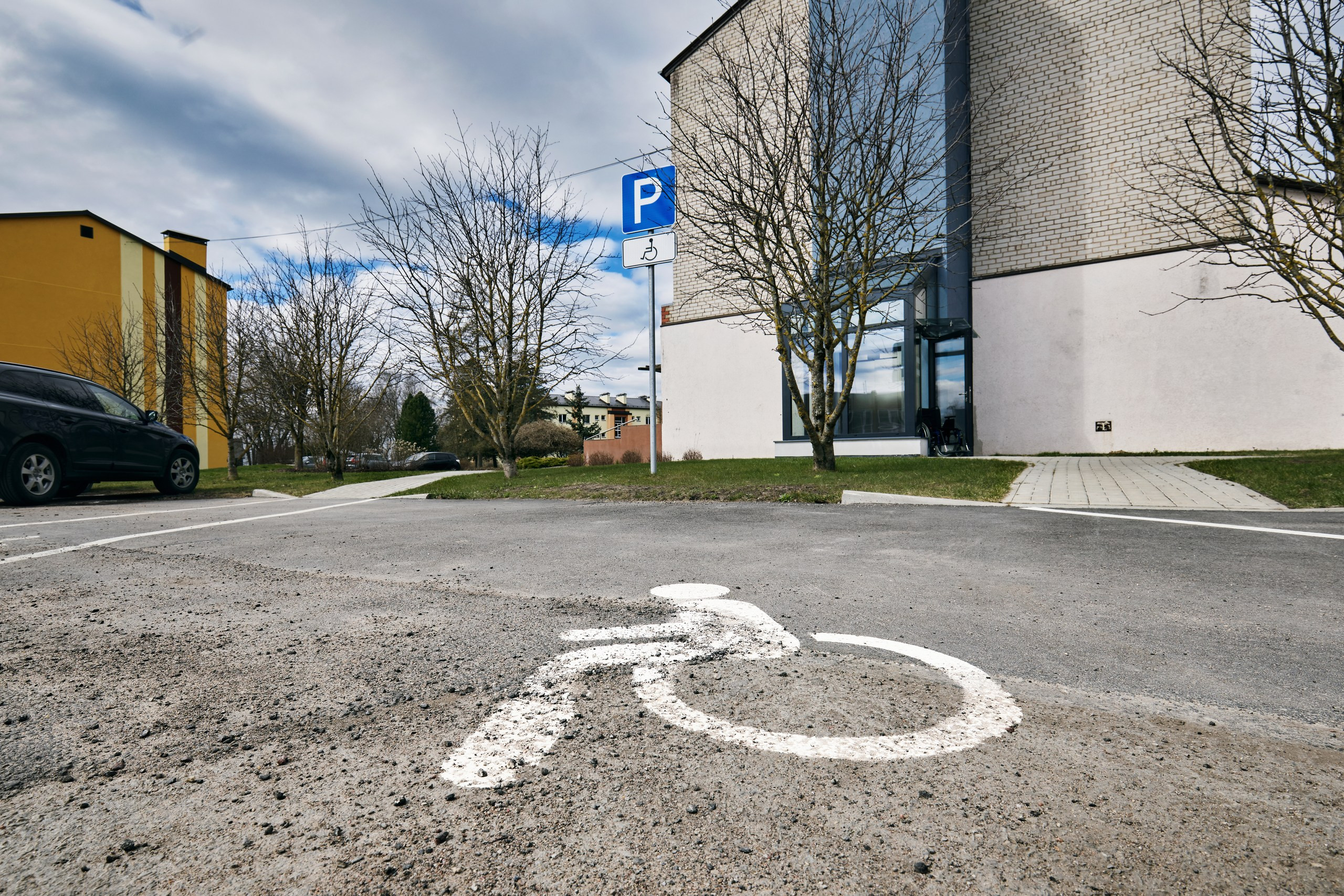 Ēkas ārpuse. Skats no ārpagalma cilvēku ar invaliditati stāvlaukuma uz stiklotu ieeju ēkā.