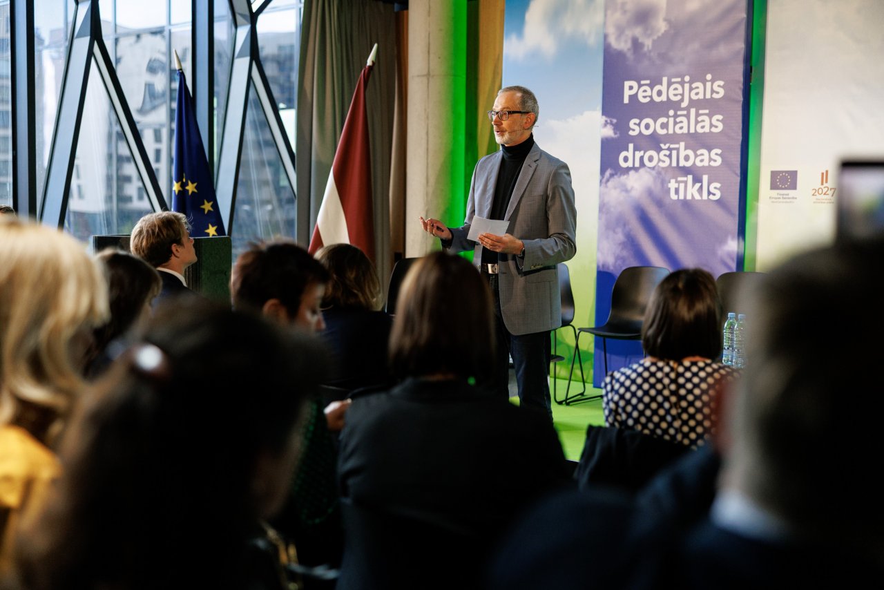 Lietišķi modernā dizainiskā konferenču telpā vīrietis ap 50 gadiem stāv priekšā auditorijai
