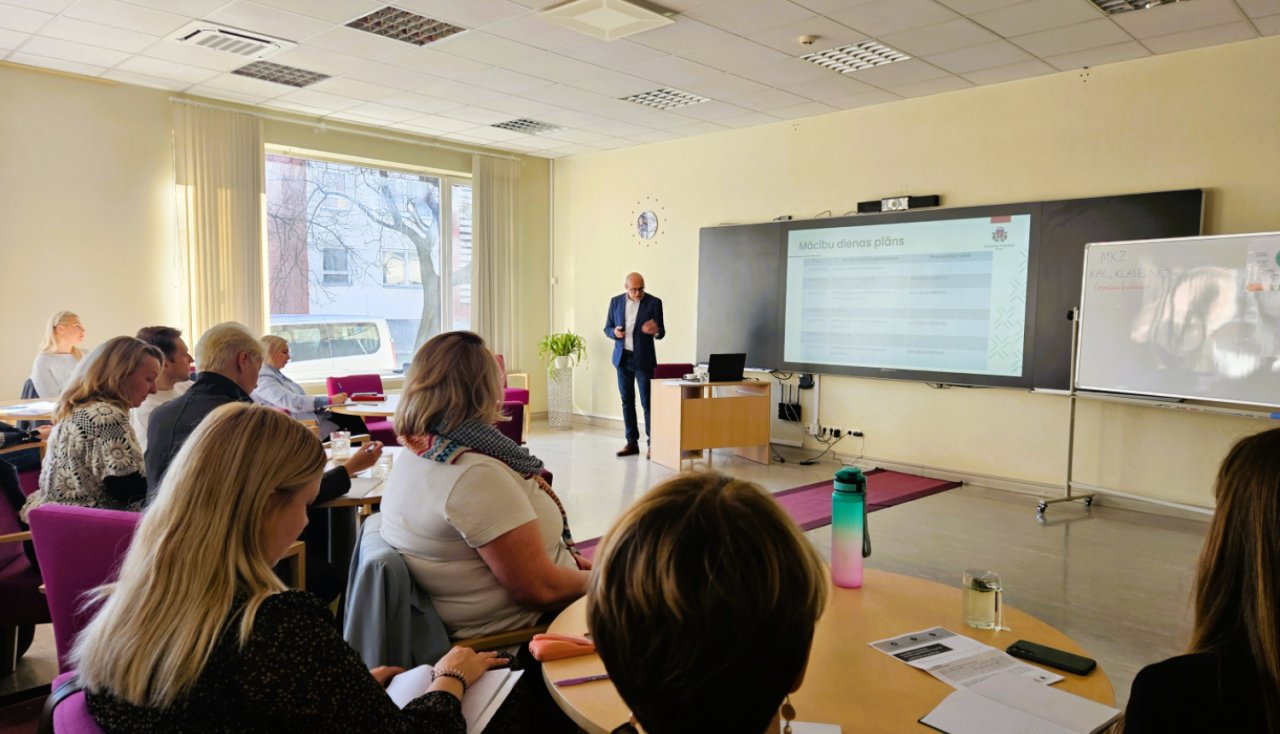 lekciju telpa. Priekšā ekrāns un pasniedzējs. Zālē sēž pusmūža klausītāji.