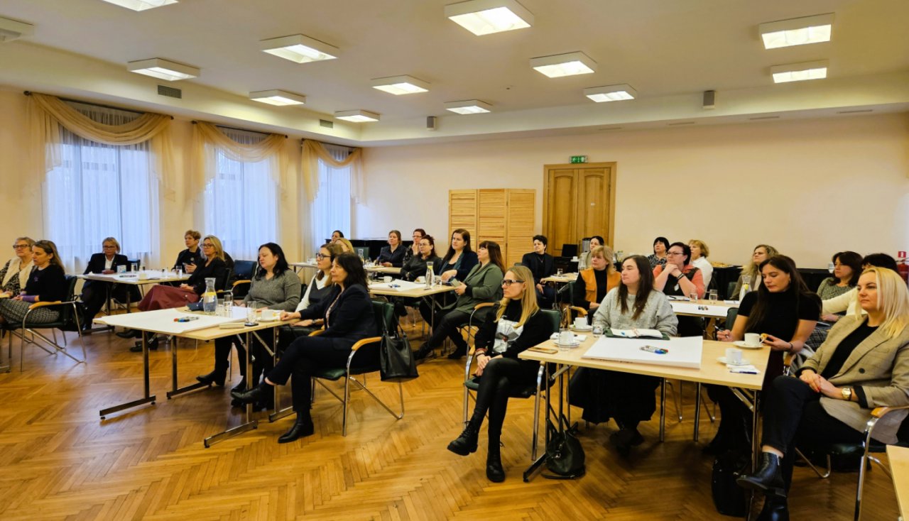 telpa ar vairākiem biroja galdiem un aptuveni 50 sēdošiem cilvēkiem, kuri klausās lekciju