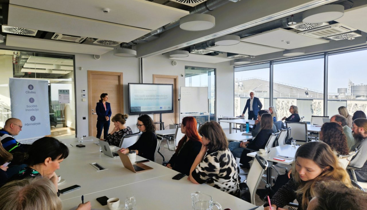 lekciju telpa ar lieliem logiem. Cilvēki randomā klausās lektoru un vietām pieraksta kaut ko datoros