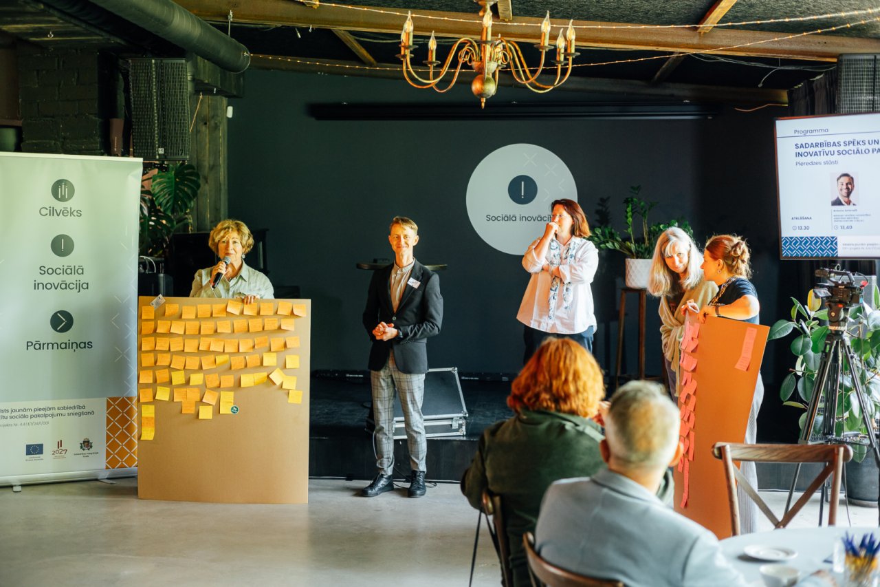 Neliela pasākuma skatuve ar vairākiem cilvēkiem uz tās. Sieviete gados runā runu