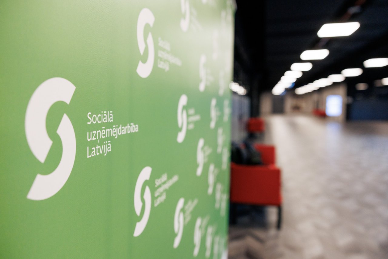 Tukšs, plašs, dizainiski glīts koridoris un kreisajā pusē logo siena zaļā krāsā. Uz sienas redzami daudz vienādi logotipi 