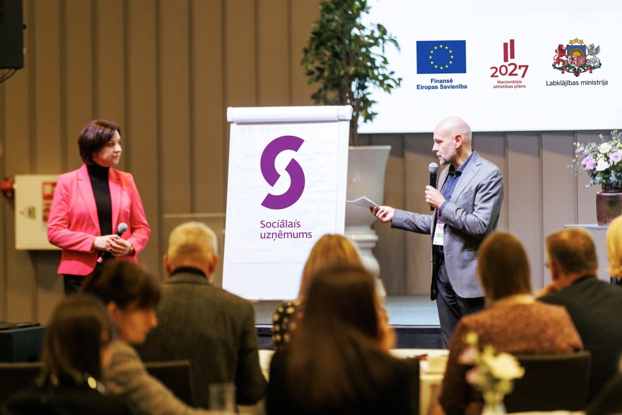 Vīrietis ar sievieti kaut ko stāsta auditorijai zālē. Fonā uzraksts 