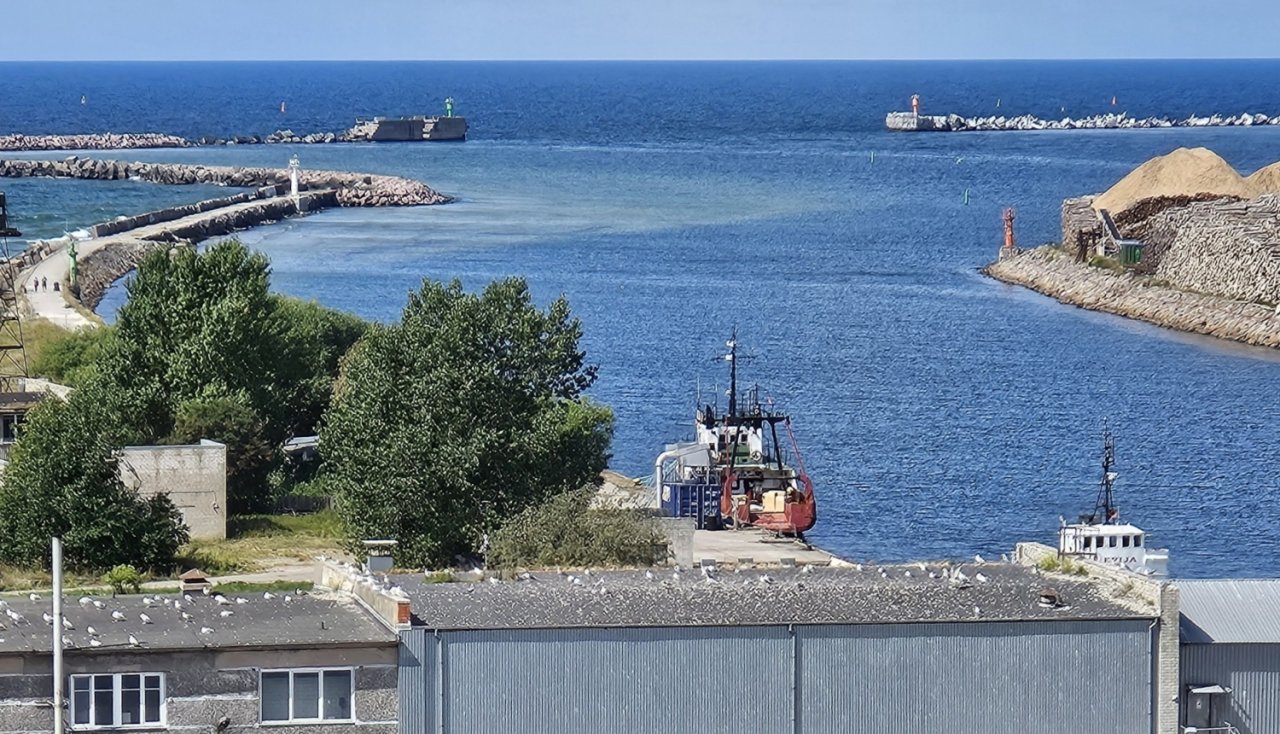 Skats uz ostas iebrauktuvi upes grīvā ar moliem tālumā un ēkām un zvejas kuģi priekšplānā