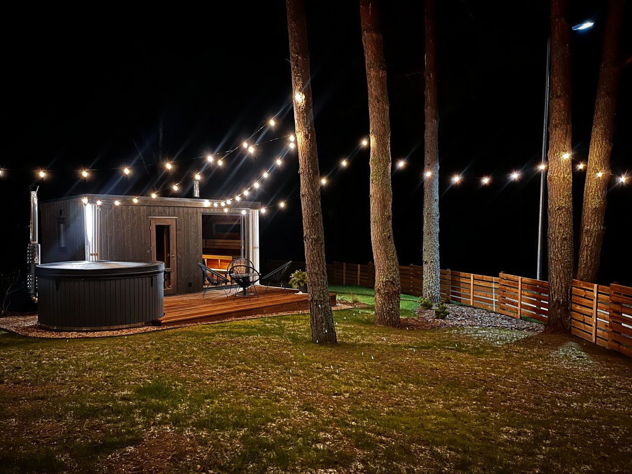 Namiņš naktī starp priedēm ar lampiņu virteni