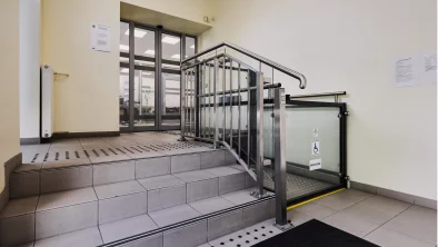 Kāpņu telpa. Vienā pusē kāpnes ar taktilajām vadulām, otrā elektriskais pacēlājs.