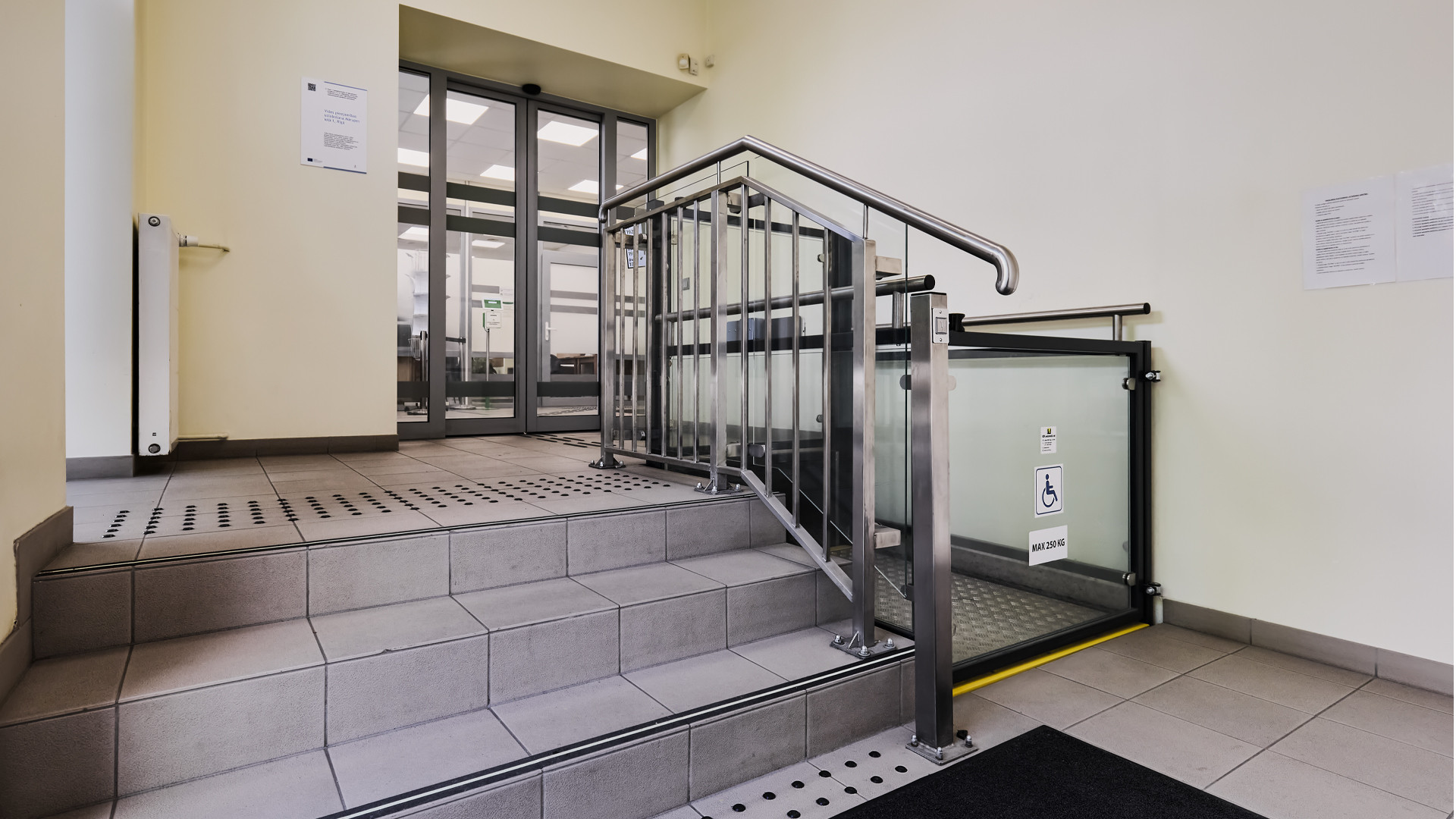 Kāpņu telpa. Vienā pusē kāpnes ar taktilajām vadulām, otrā elektriskais pacēlājs.