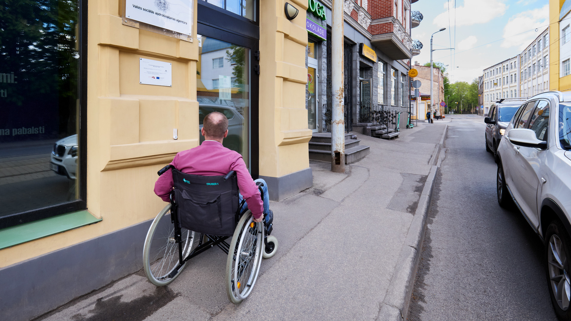 Cilvēks ratiņkrēslā uz asfaltētas ietves pie dzeltenas mūra mājas ieejas.