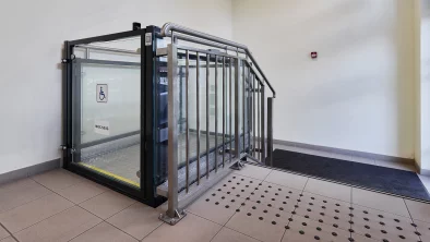 Gaiša kāpņu telpa. Labajā pusē kāpnes ar taktilo virsmu. Kreisajā pusē elektriskais pacēlājs cilvēkiem ar invaliditāti