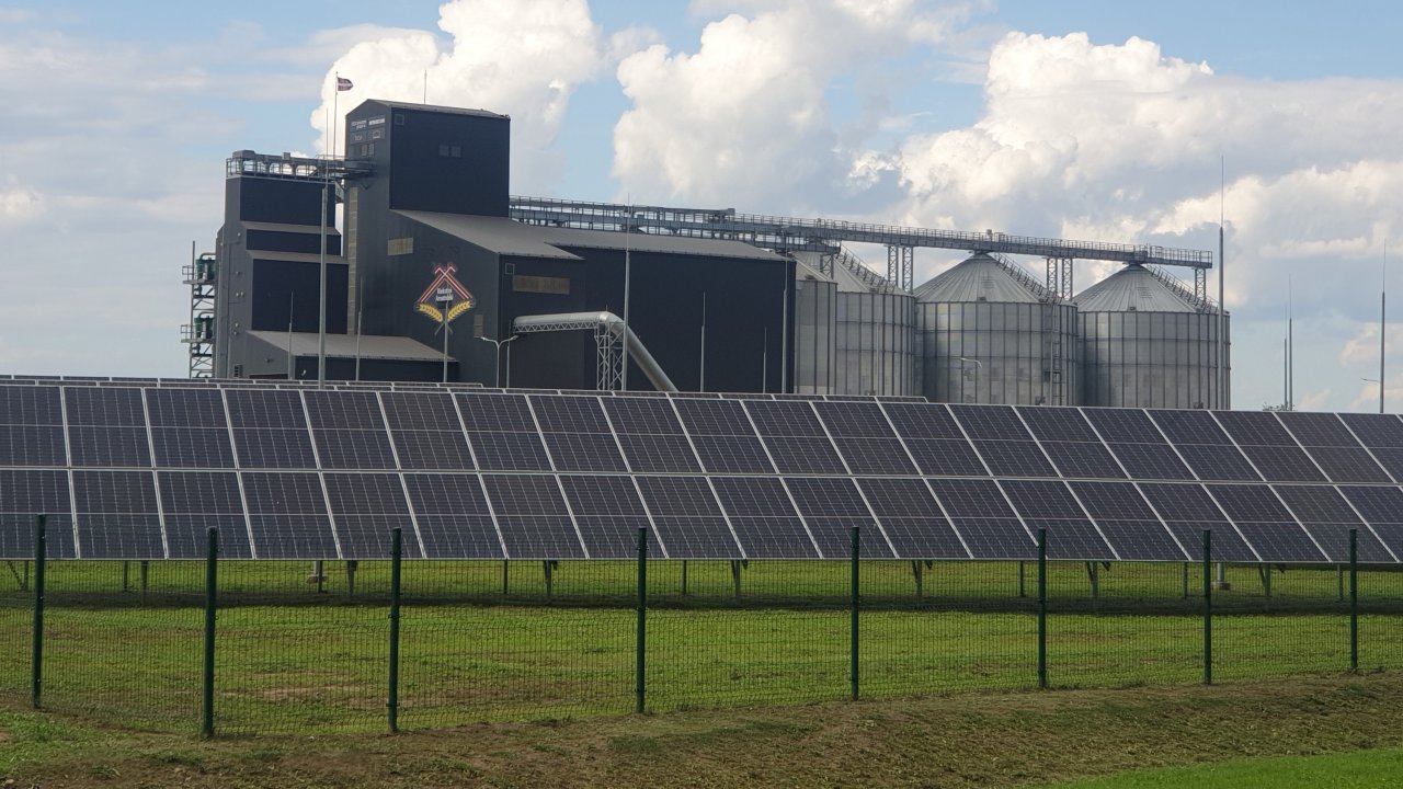 priekšplānā saules paneļu rinda, aiz tiem graudu kalte