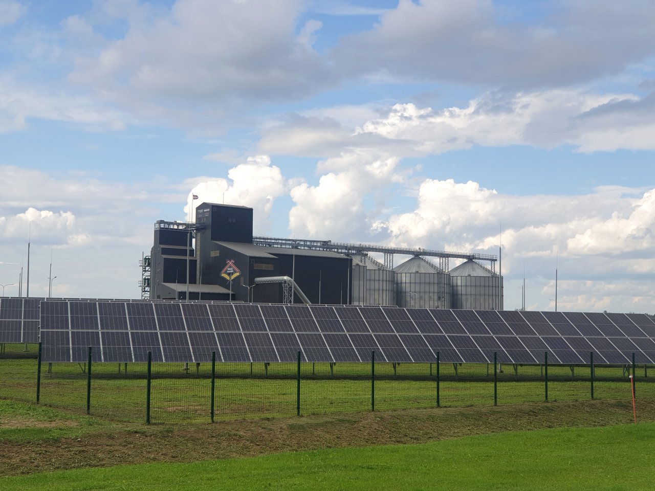 zaļā laukā saules paneļu rinda, aiz tās graudu uzglabāšanas torņi un kaltes ēka
