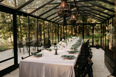 Siltumnīca, iekārtota svinībām no iekšpuses ar garu galdu pa vidu, kuram uzklāts balsts galdauts un salikti trauki