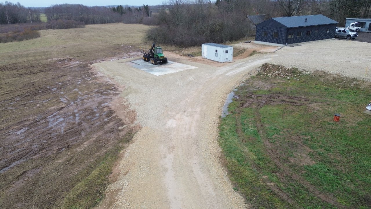 ZS Laurii šņembots laukums tehnikas novietošanai, kurā stāv zaļš traktors, tālākajā stūrī melna garāža. bildēts no drona